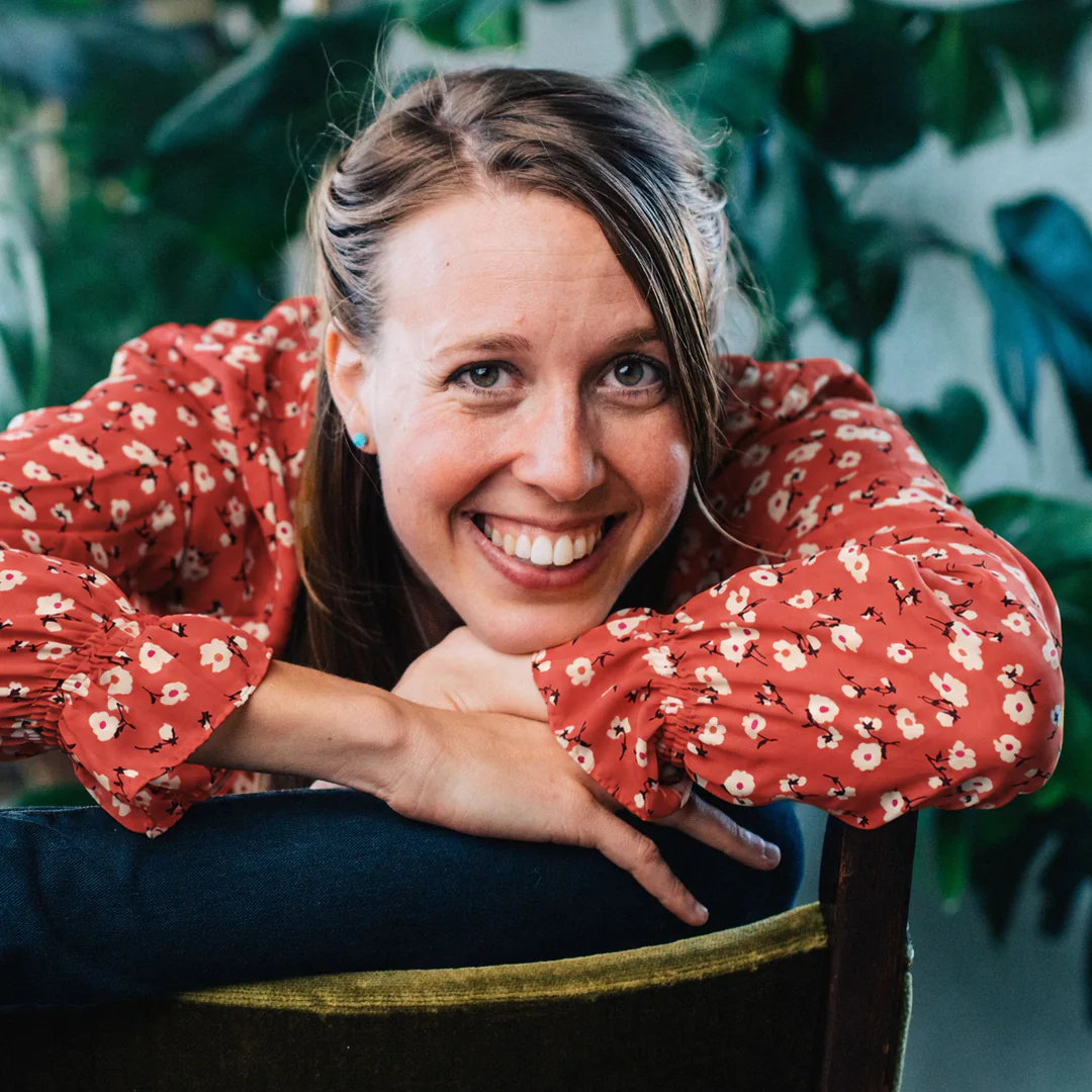 Hailey Jones laughing in a coral floral blouse, leaning on the back of a chair in front of green plants