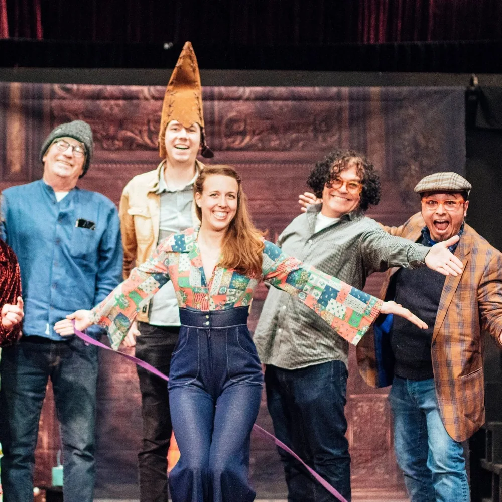 Hailey Jones in a patchwork shirt takes a bow on stage with five costumed cast members, one in a tall pointed felt hat