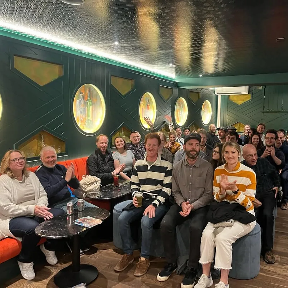 A full house of about 40 guests on couches and stools, laughing and clapping at a comedy show