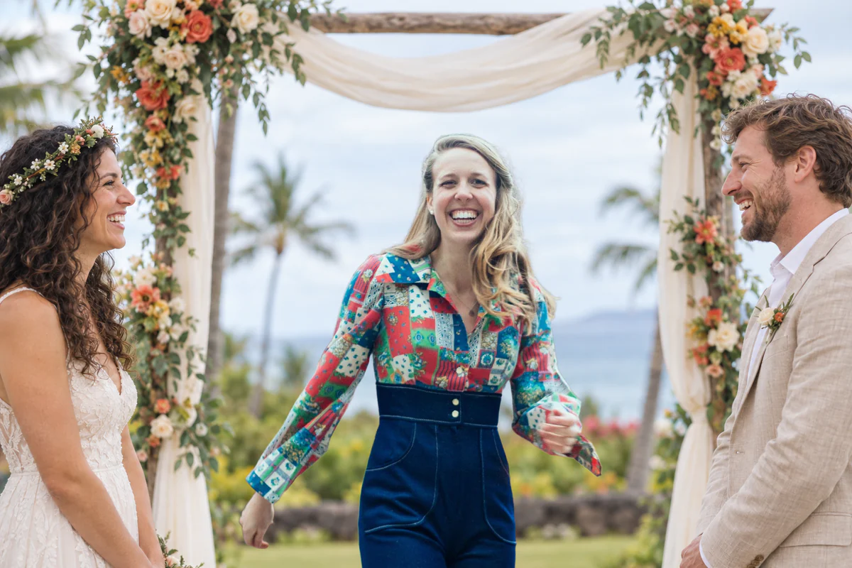Hailey Jones laughing as she officiates an outdoor wedding under a flower arch, the couple facing each other on either side of her