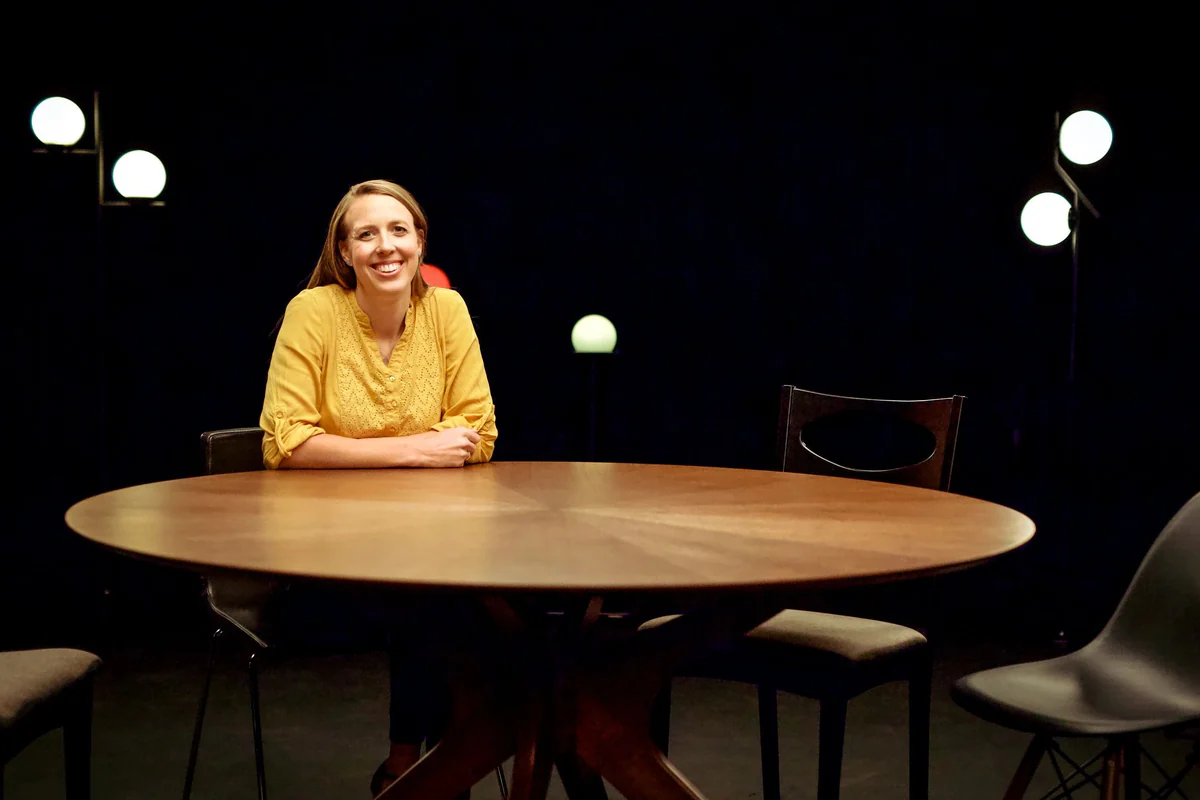 Hailey Jones smiling at a round wooden dinner table under globe lamps with empty chairs around it